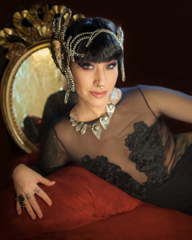 Press photo, portrait: Sharon Kihara reclining on a red cushion in front of a gold-framed chair, wearing an ornate headpiece