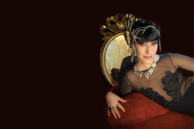 Press photo, landscape: sharon Kihara reclining against a gold-framed chair, wearing an ornate gold headpiece and a sheer black lace top